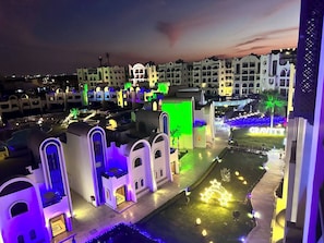 A hotel with multiple buildings, each featuring arched windows and balconies. The buildings are illuminated with various colors, and there is a well-maintained lawn area with decorative lighting. - Luxury Apartment- Sahl Hasheesh- Gravity Hotel (Sahl Hasheeh)