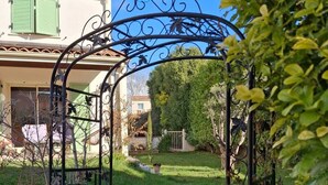 A garden with a metal archway leading to a house with green shutters. - Beautiful Mediterranean villa with large pool ideal for families (Villeveyrac)