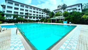 A swimming pool with a tiled deck, surrounded by a hotel building with multiple balconies and a covered area with lounge chairs. - Spacious Corner 1-bedroom Condo With 2 Balconies - Baan Suan (Pattaya)