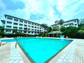 A swimming pool with a tiled deck, surrounded by a hotel building with multiple balconies and a covered area with lounge chairs. - Spacious Corner 1-bedroom Condo With 2 Balconies - Baan Suan (Pattaya)