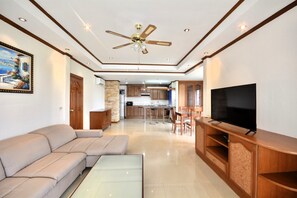 A modern living room with a sofa, a glass coffee table, a TV on a wooden stand, and a ceiling fan. - Spacious Corner 1-bedroom Condo With 2 Balconies - Baan Suan (Pattaya)