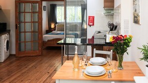 Comedor con una mesa para dos, cocina al fondo y dormitorio con cama y ventana.