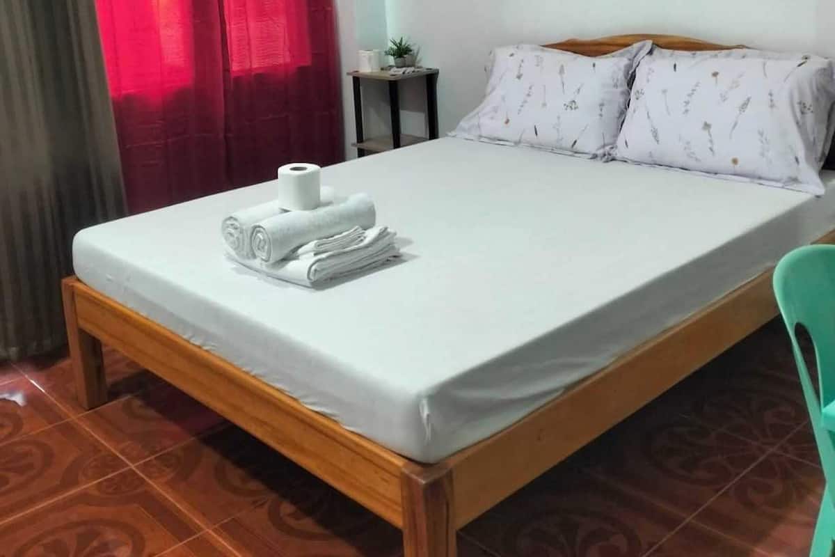 Cama bem arrumada com lençol branco, toalhas enroladas e um rolo de papel higiênico no apoio para os pés.