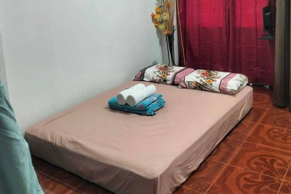 Uma cama pequena e bem arrumada, com um edredom rosa e duas almofadas decorativas, uma pequena mesa com toalhas enroladas, e um vaso com flores.