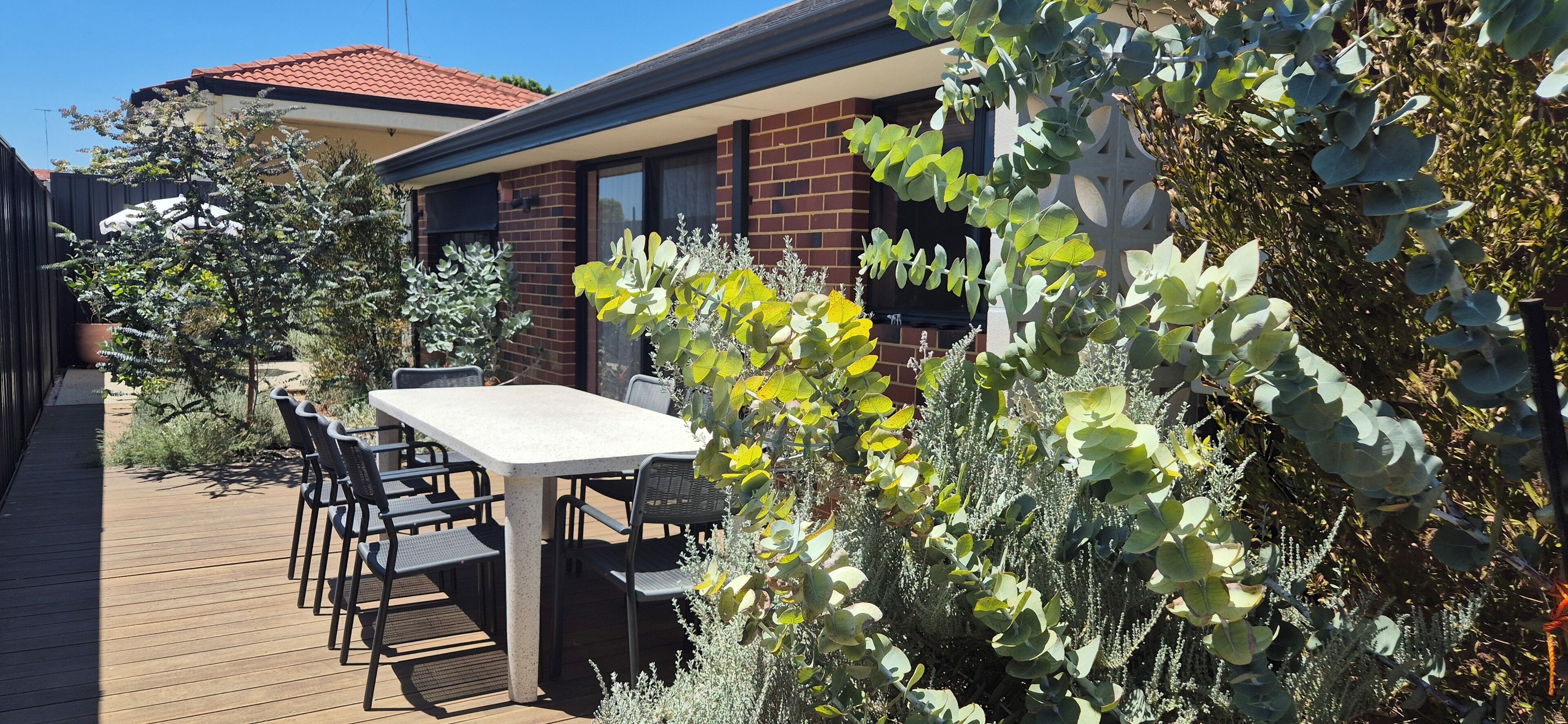 A patio with a table and chairs, surrounded by various plants and a brick building in the background.