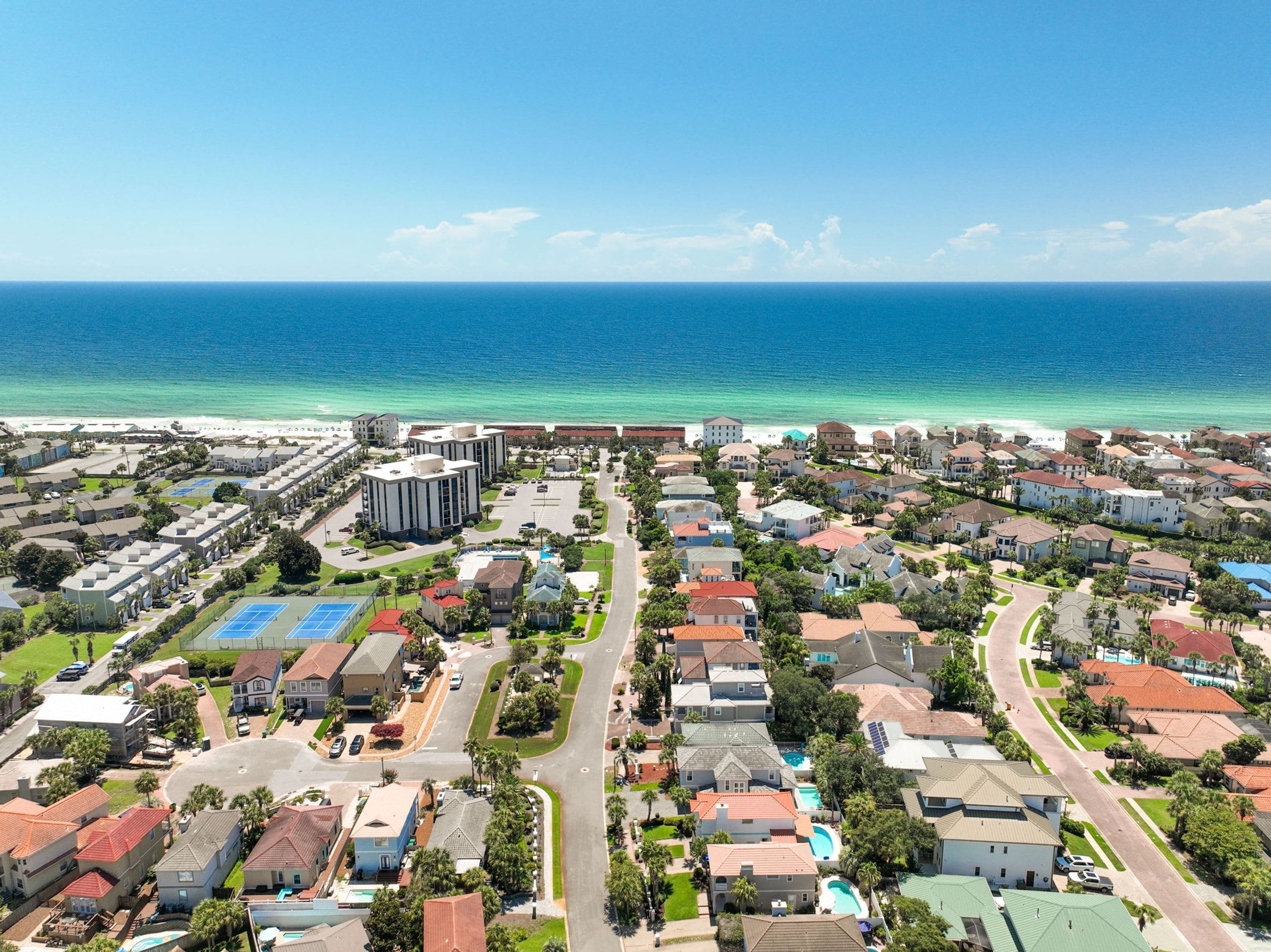 Vue aérienne d’un quartier résidentiel situé sur la côte, avec des maisons, un parking et une plage en arrière-plan.