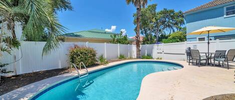 A swimming pool with a curved edge, surrounded by a white fence, palm trees, and a blue house in the background.