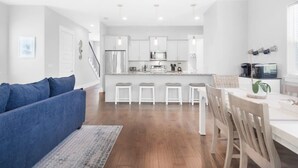 A modern kitchen with white cabinets, a central island with stools, and a dining area with a white table and chairs. - Cabin by the Sea - 3bdr - Pool Walk 2 Beach (Miramar Beach)