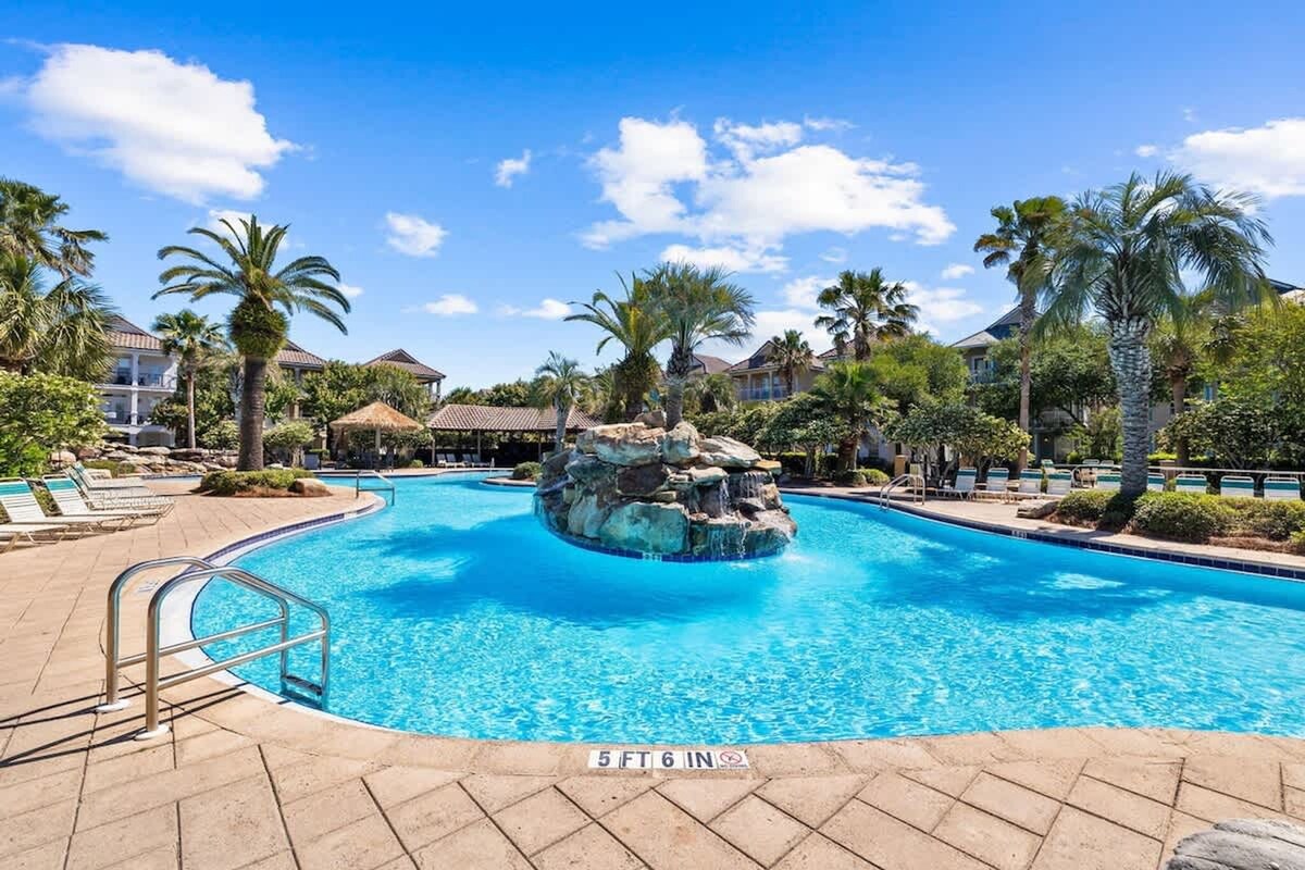 Piscina com um elemento rochoso, rodeada por palmeiras e cadeiras de descanso.