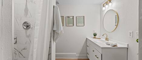 A bathroom with a shower, a sink with a gold faucet, and a mirror with lights. There are framed pictures on the wall and a towel hanging on the door.