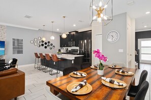 A modern kitchen with dark cabinets, a central island, and a dining area with a wooden table set for four. - Super Swank - Aesthetic Designs Amenities (Davenport)