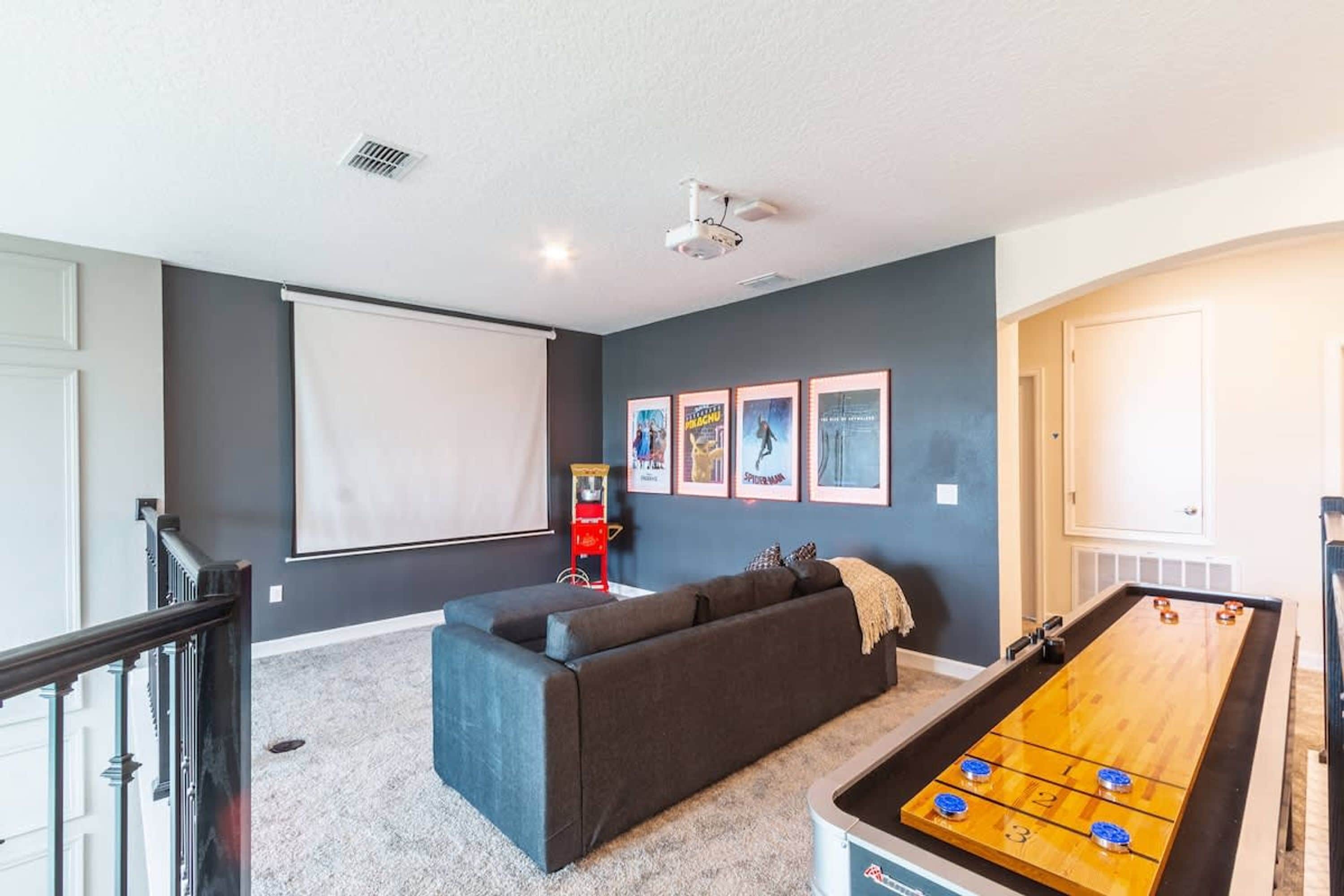 Una habitación con un sofá de color oscuro, una mesa de fútbol de mesa, una pantalla de proyector y carteles de películas enmarcados en la pared.