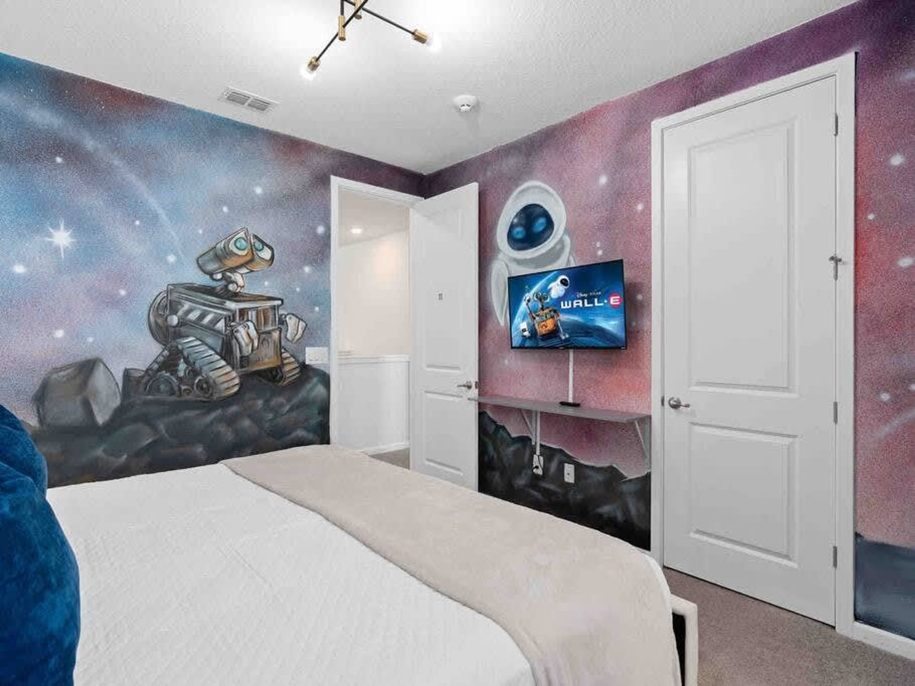Een slaapkamer met een muurschildering over het thema ruimte, een bed, een televisie op een standaard en twee witte deuren.
