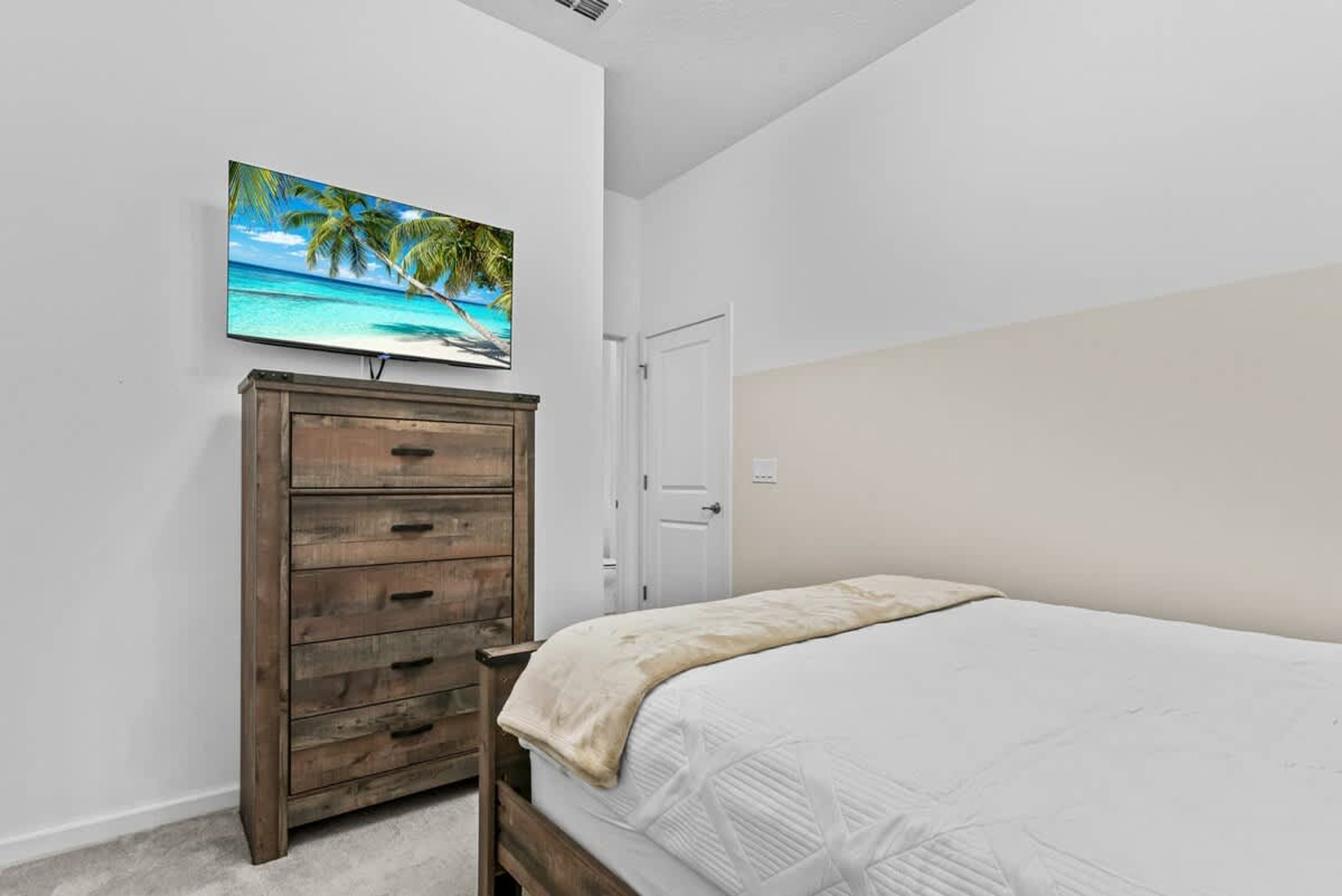 A bedroom with a bed, a wooden dresser, a television mounted on the wall, and a door.