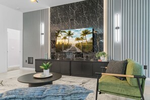 Una sala de estar moderna con un televisor de pantalla plana grande montado en una pared de mármol, un mueble de madera oscura debajo, una mesa redonda con una planta y un sillón verde.
