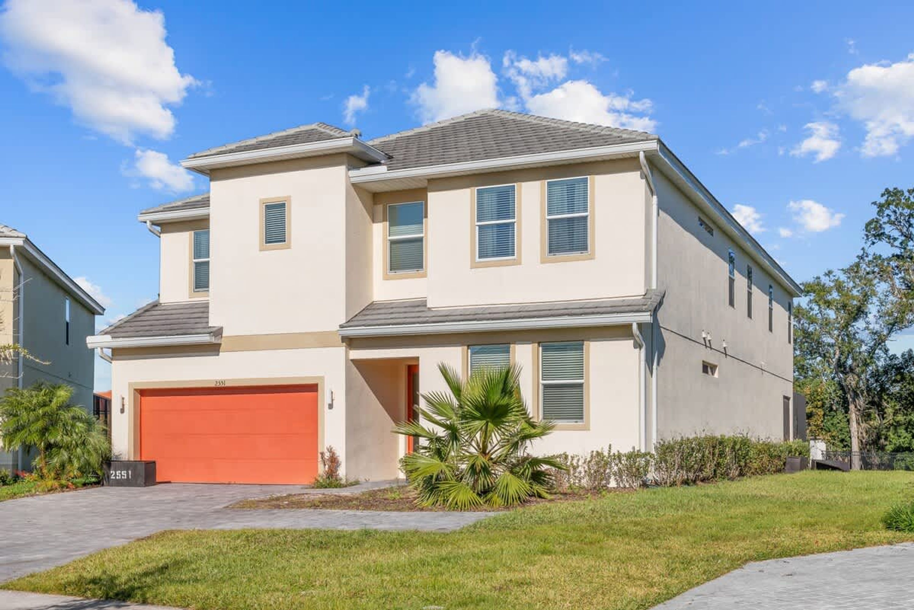 A two-story house with a red garage door, a small front yard with a palm tree, and a clear blue sky.