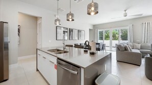 A modern kitchen with a central island, stainless steel appliances, pendant lighting, and a dining area with a sofa and table. - Resort-style 4-bed Loft Condo (Kissimmee)
