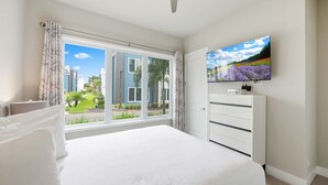A bedroom with a large bed, a TV mounted on the wall, and a window with a view of buildings and greenery. - Resort-style 4-bed Loft Condo (Kissimmee)