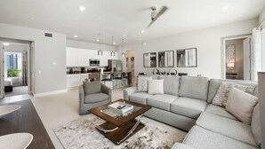 A modern living room with a grey sofa, a coffee table, and a dining area with a kitchen in the background. - Resort-style 4-bed Loft Condo (Kissimmee)