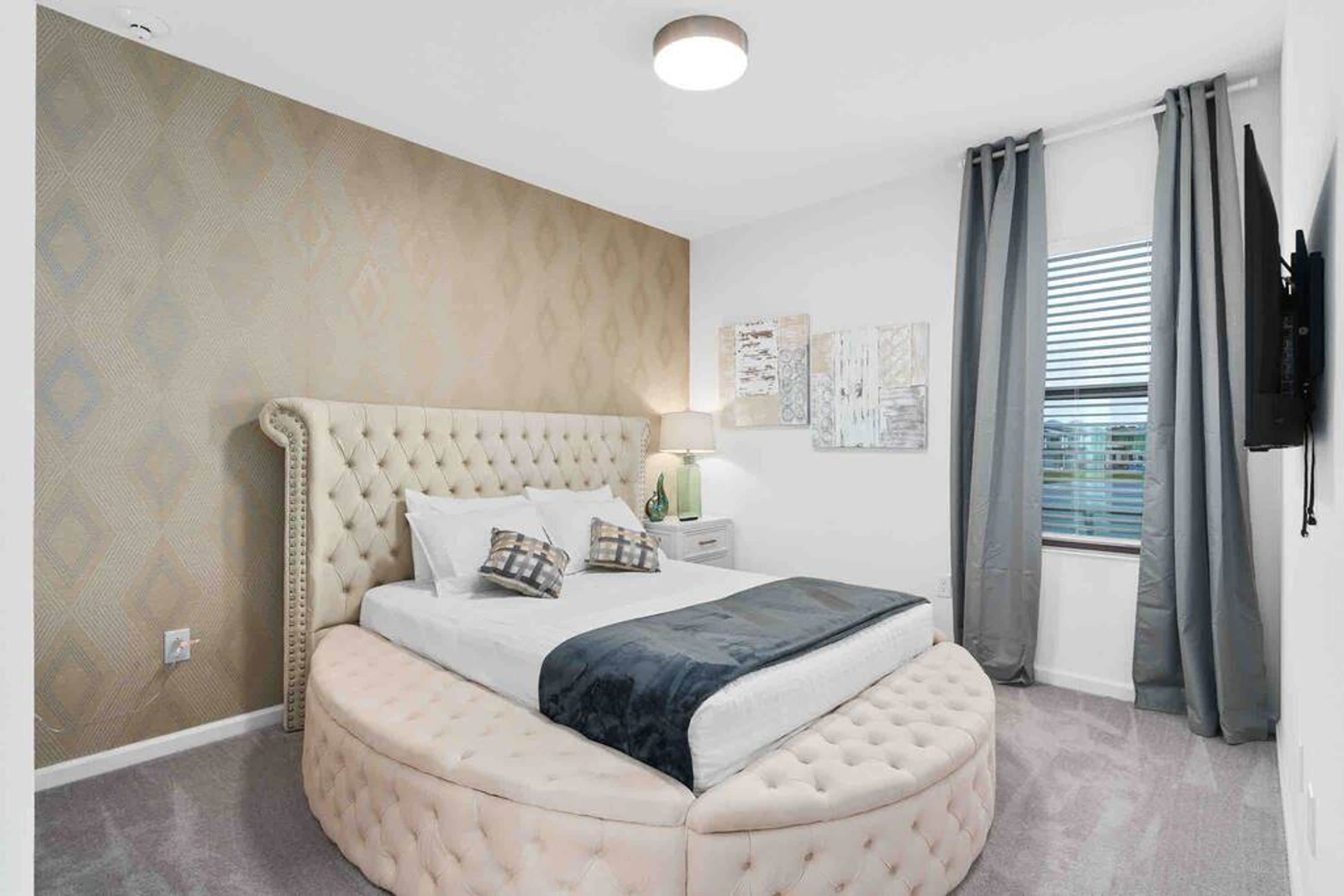 A bedroom with a tufted headboard, a round ottoman, a bed with pillows, a nightstand with a lamp, and a flat-screen TV mounted on the wall.