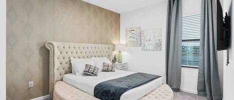 A bedroom with a tufted headboard, a round ottoman, a bed with pillows, a nightstand with a lamp, and a flat-screen TV mounted on the wall.