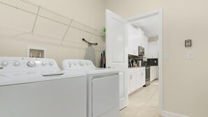 Une buanderie équipée de deux machines à laver et deux sèche-linge à chargement frontal, d’un étendoir et d’un coin cuisine visible à travers une porte ouverte.