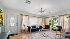 Un salón amplio con un sofá azul oscuro, una mesa de centro y sillones negros. Se ve una puerta con un panel de vidrio decorativo y vistas a plantas afuera.