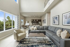 Un salon spacieux avec un canapé d’angle gris, un fauteuil beige et une table basse. Une grande fenêtre offre une vue sur la verdure environnante.