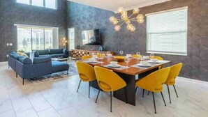 A modern living room with a sofa, dining table, and chairs, a television, and a large window. - Pool Spa Arcade Games - 5 min Walk to Water Park (Kissimmee)