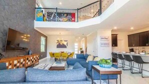 A spacious living area with a large sofa set, a dining table with yellow chairs, a kitchen area, and a balcony with a railing. - Pool Spa Arcade Games - 5 min Walk to Water Park (Kissimmee)