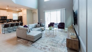 A spacious living area with a large sofa, a coffee table, and a TV mounted on the wall. There is a kitchen in the background with modern appliances and bar stools. - Ultimate Comfort Prime Location Luxury Villa (Kissimmee)