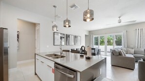 Eine moderne Küche mit Kochinsel, Edelstahlgeräten, Pendelleuchten und Blick in den Wohnbereich mit einer Couch.