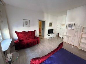 Ein modernes Zimmer mit einem roten Sofa, einem blauen Bett, einem Fernseher und einem weißen Schreibtisch.