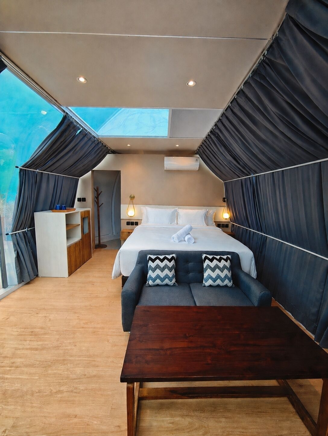 A modern bedroom with a large bed, a sofa, a wooden table, and a skylight.