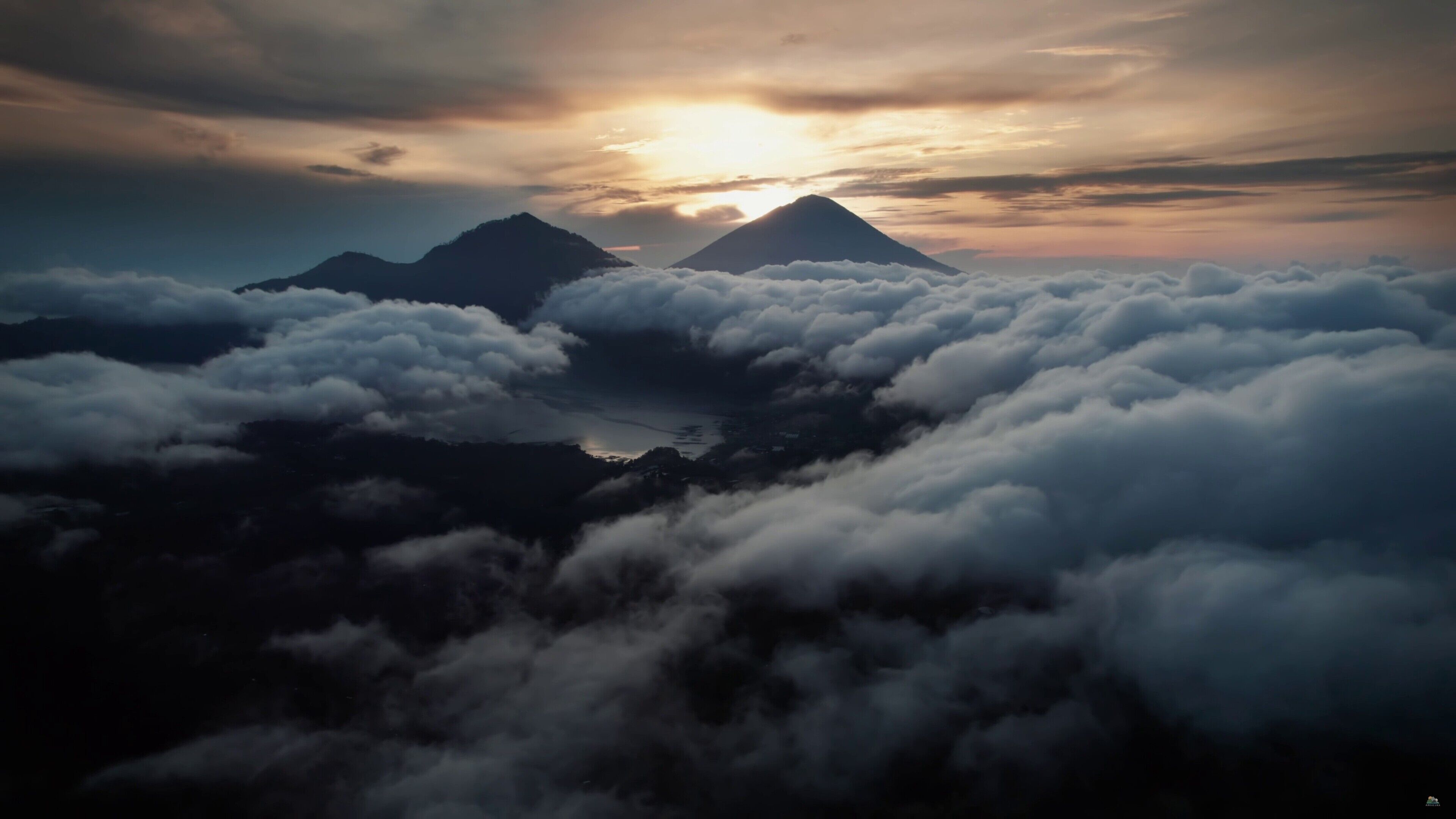 Paisaje montañoso con nubes abajo y un sol poniéndose al fondo.