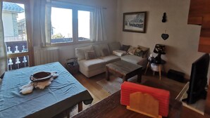 Una sala de estar acogedora con un sofá, una mesa de centro y una mesa de comedor con un mantel individual. Se aprecia una ventana con cortinas, un cuadro enmarcado en la pared y una mesita con un adorno.