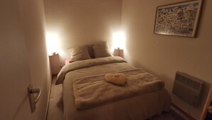 Un dormitorio pequeño y acogedor con cama, lámparas de noche, un radiador y un cuadro en la pared.