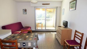 A small, well-lit room with a sofa, a table with a quilt, a TV on a wooden cabinet, and a sliding glass door leading to a balcony. - Apartment in Pin Rolland by Ste Asile Beach (Saint-Mandrier-sur-Mer)