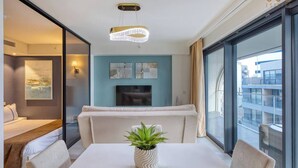 Una habitación de hotel moderna con un ventanal grande, un televisor de pantalla plana, un sofá, una mesa de comedor con sillas y una planta en maceta.