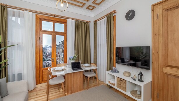 A room with a wooden floor, a dining table with chairs, a TV on the wall, and a window with curtains. - Acme Studio 2 (İstanbul)