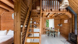 Interior d'una cabana de fusta amb un dormitori a la llosa, una zona de dinar i un foc de llenya.