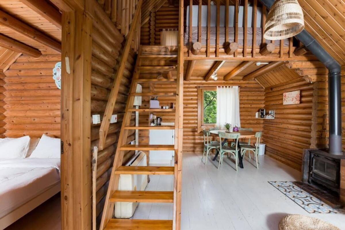 A log cabin interior with a loft bedroom, a dining area, and a wood-burning stove.