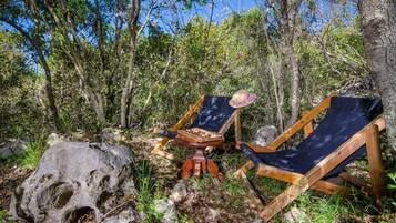 Deux fauteuils en bois avec des coussins, un grand rocher et un tabouret en bois, le tout situé dans une zone boisée.
