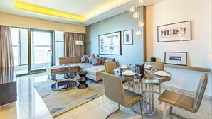 A modern living room with a sofa, dining table, and chairs. There are framed pictures on the wall and a window with curtains. - Superhost 1BR DAMAC Towers Paramount (Dubai)