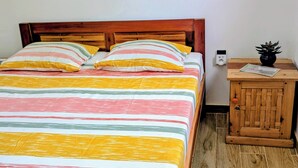 A bed with a striped bedspread and two pillows, a wooden nightstand with a plant and a card, and a wall socket.