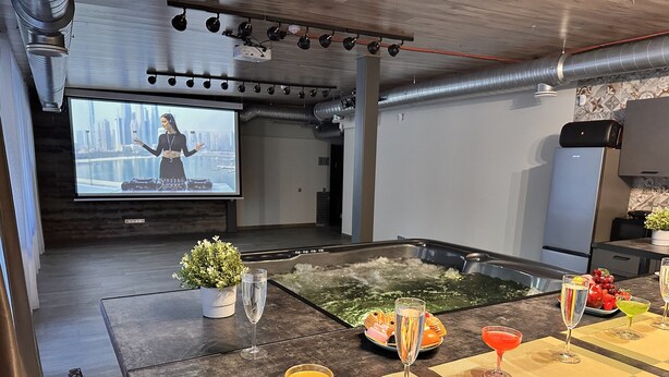 A modern bar area with a heated floor, a large screen displaying a presenter, and a variety of drinks on the counter.