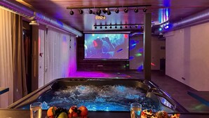 A spa area with a hot tub, a large screen displaying a concert, and a variety of fruits and drinks on the table.