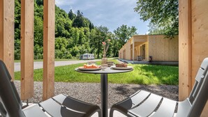 A wooden structure with a table and two chairs in front, a small table with food and drinks, and a forested background.