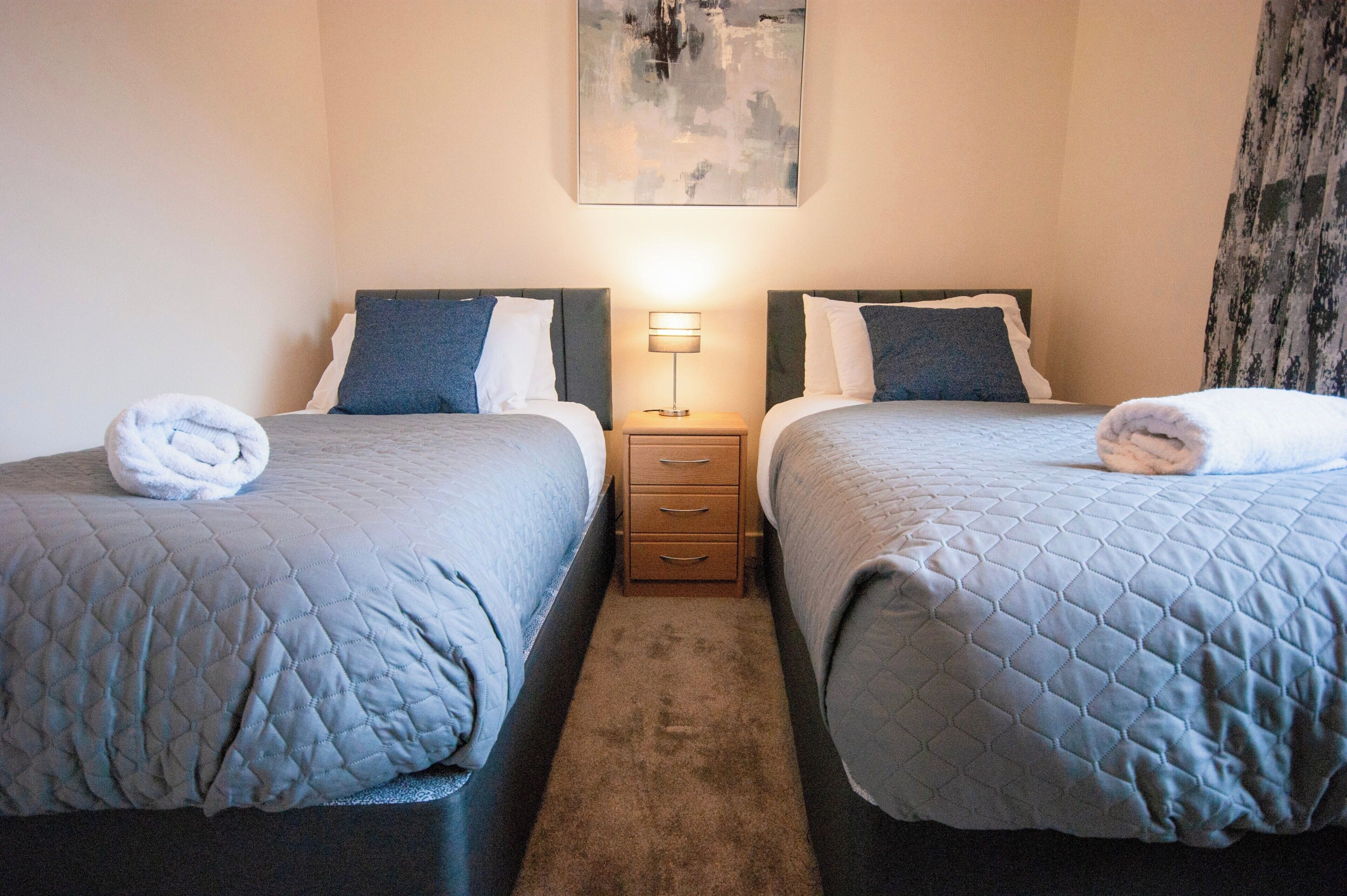 A hotel room with two beds, each with a quilted bedspread and pillows, a bedside table with a lamp, and a painting on the wall.