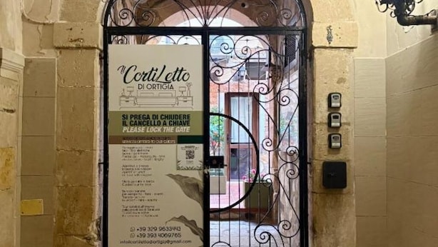A hotel entrance with a wrought iron gate, a sign for 'Corti/Corti di Portigio' with contact information, and a view of the street outside. - Il Cortiletto di Ortigia - Two-room apartment (Siracusa)